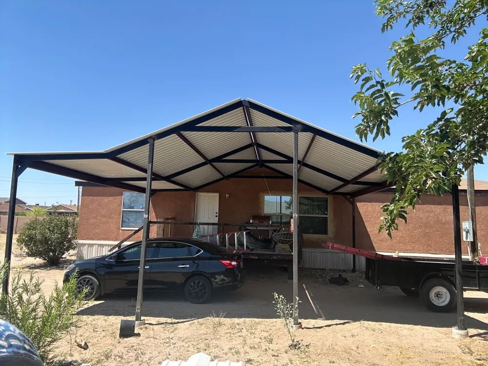 Black car and trailer parked under a metal carport beside a brick house in a desert yard.