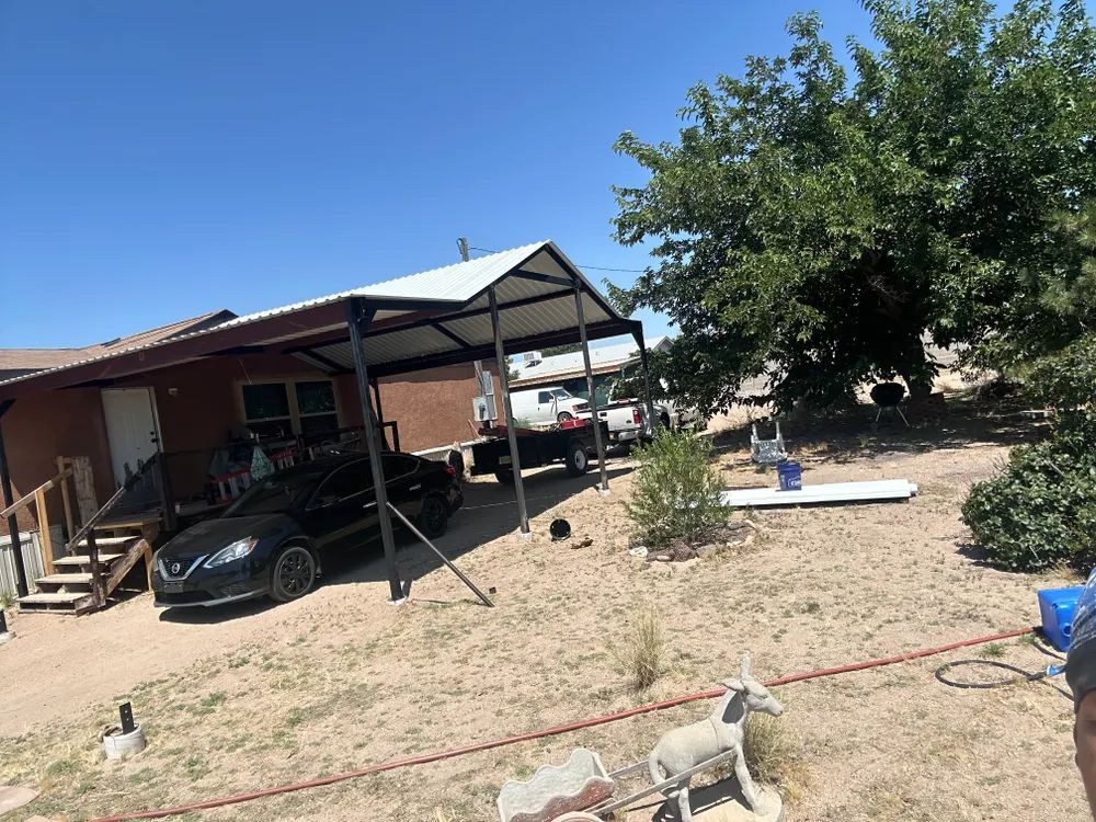 Backyard with a carport, black car, trees, and a dog in the yard under a bright blue sky
