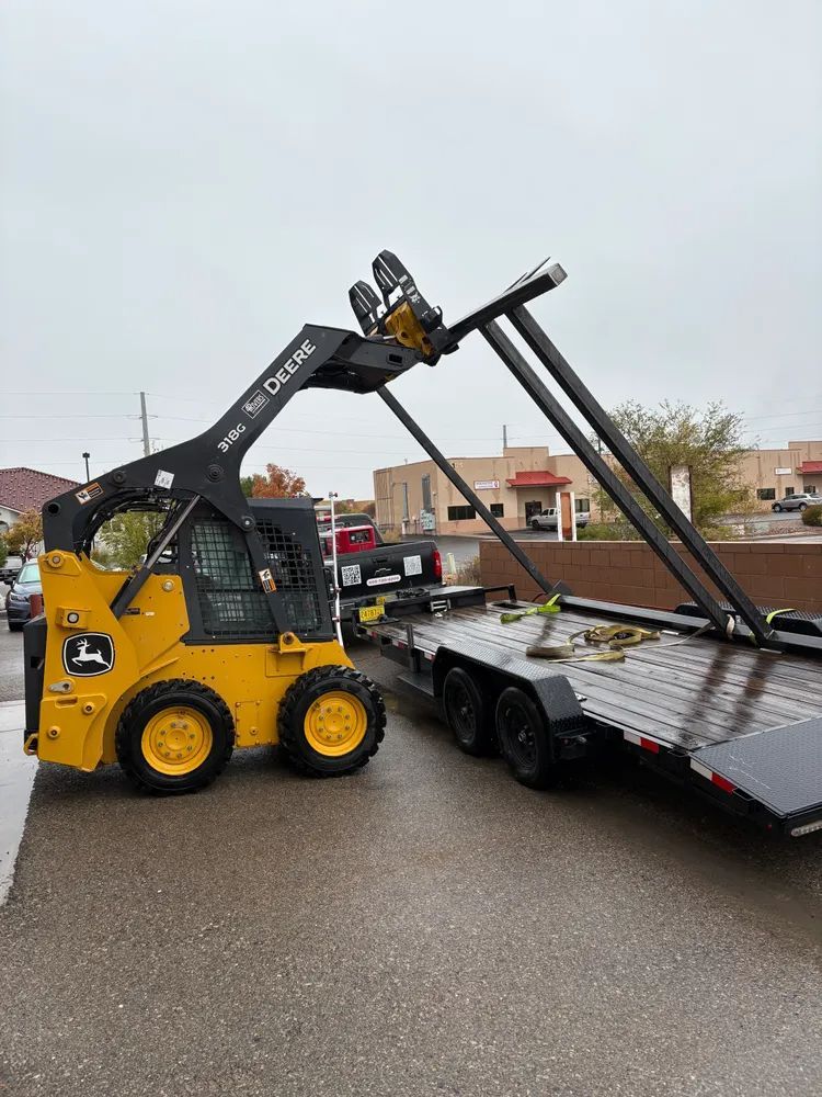 Yellow skid steer loading metal beams onto a flatbed trailer in a wet parking lot