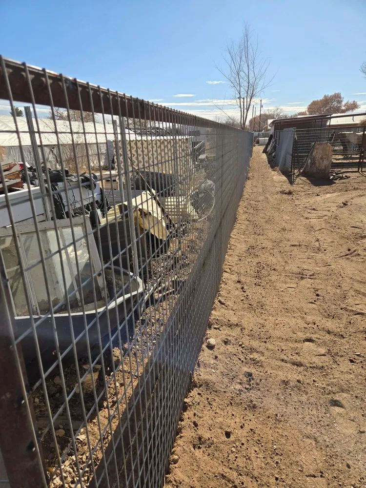 Wire fence along a dirt path beside a yard with debris and structures under a clear sky.