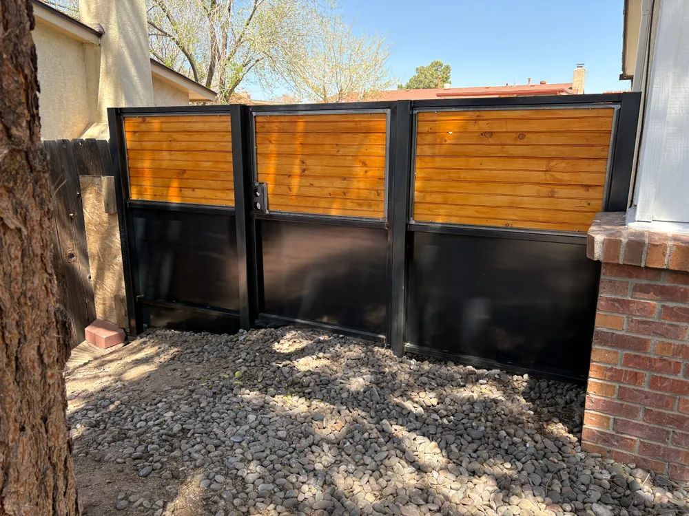 Black metal-and-wood fence panels beside a gravel yard and brick wall in daylight