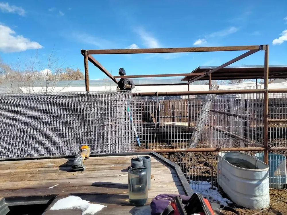 Worker atop a metal frame behind a tarp-covered fence, with tools and buckets in the foreground.