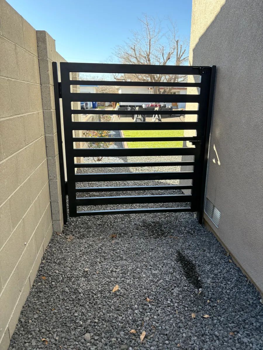 Black slatted gate between beige walls over gravel walkway, with a view to outdoors beyond.