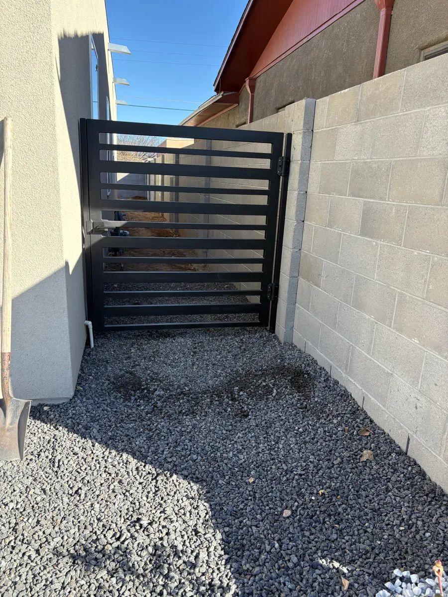 Narrow gravel driveway ending at a black metal gate between a house wall and block wall.