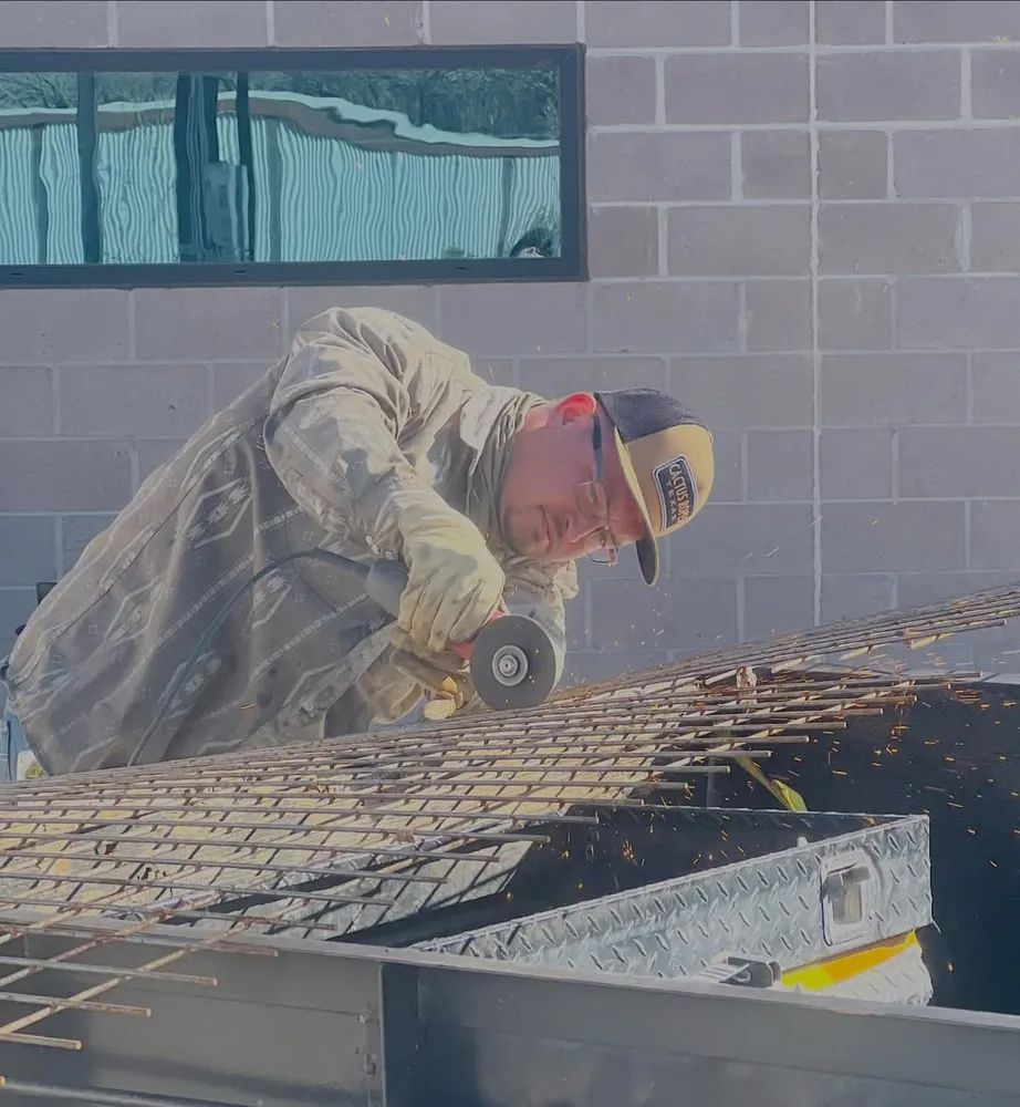 Worker in tan jacket and cap cutting rebar on a construction site beside a brick wall.