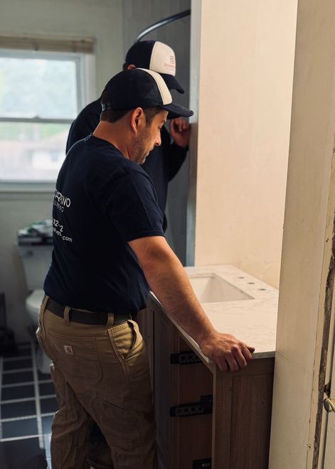 Plumber in gray overalls and gloves works under a kitchen sink with a power tool.