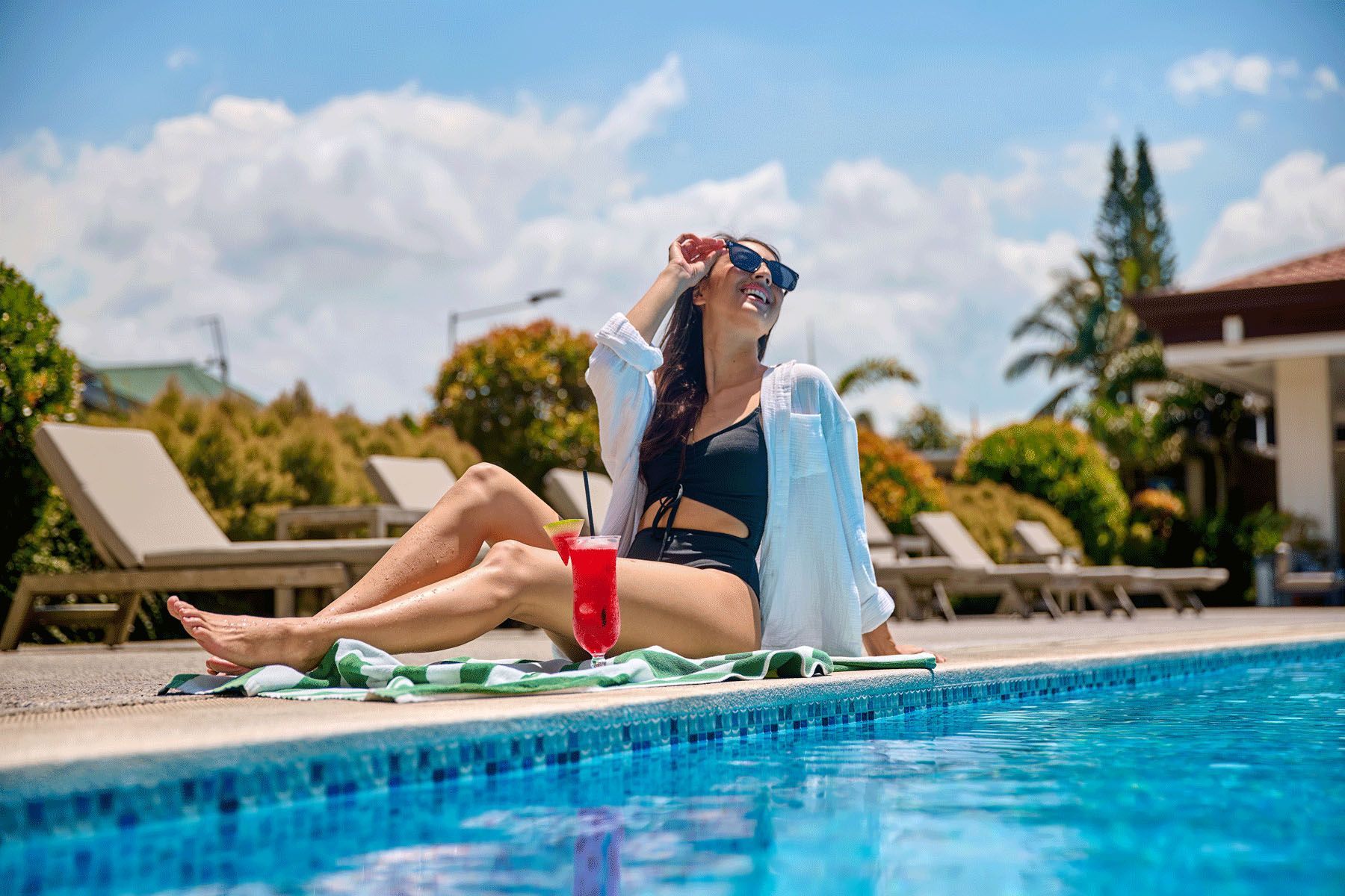 Woman in sunglasses and swimsuit relaxes by a pool, holding drink on a sunny day.