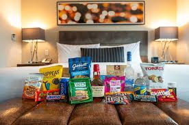 Assorted snack bags arranged on a bed in a warmly lit bedroom with bedside lamps.