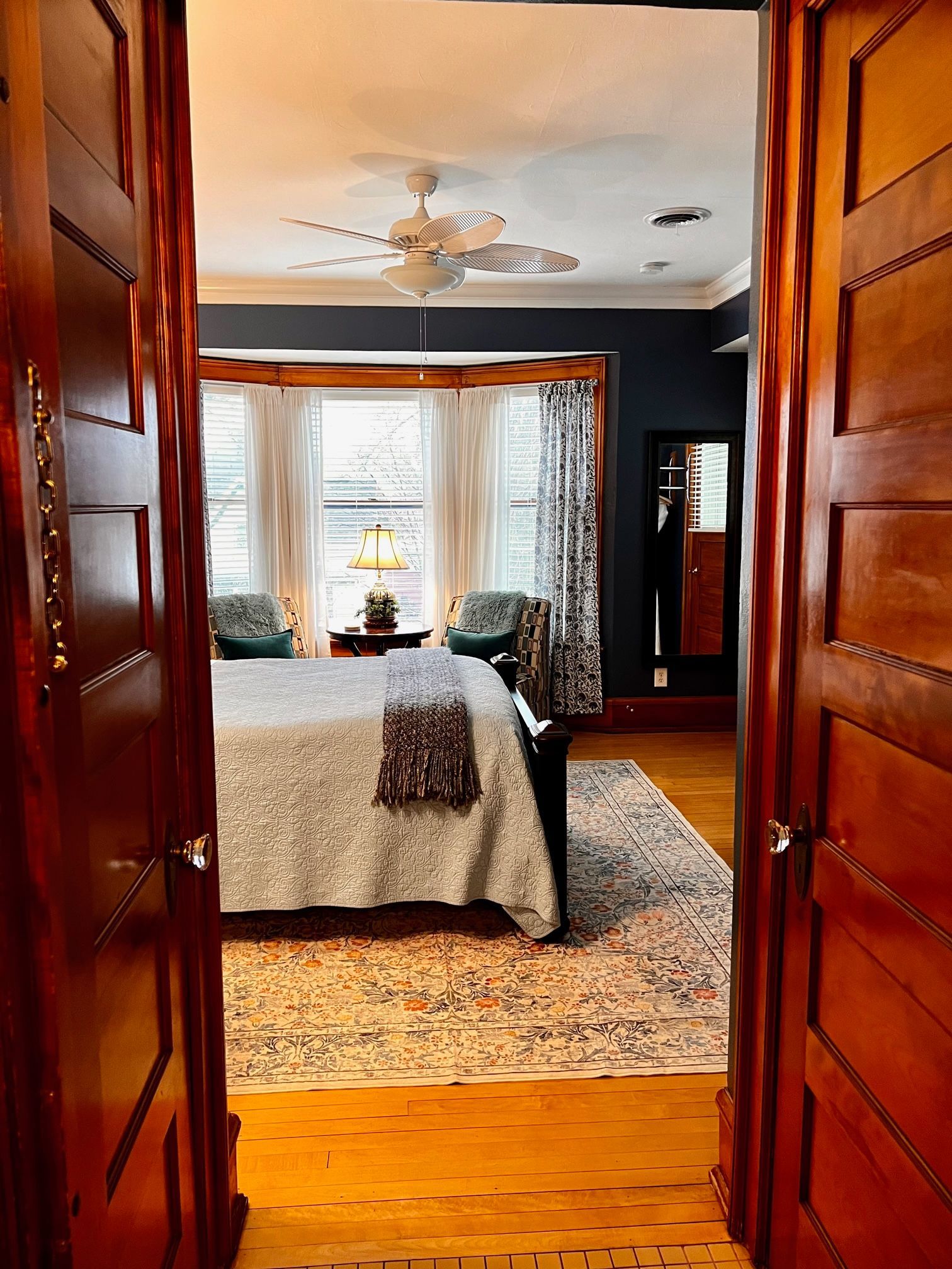View through open doorway into a bedroom with wooden floors, a bed, and window seat with a patterned rug.