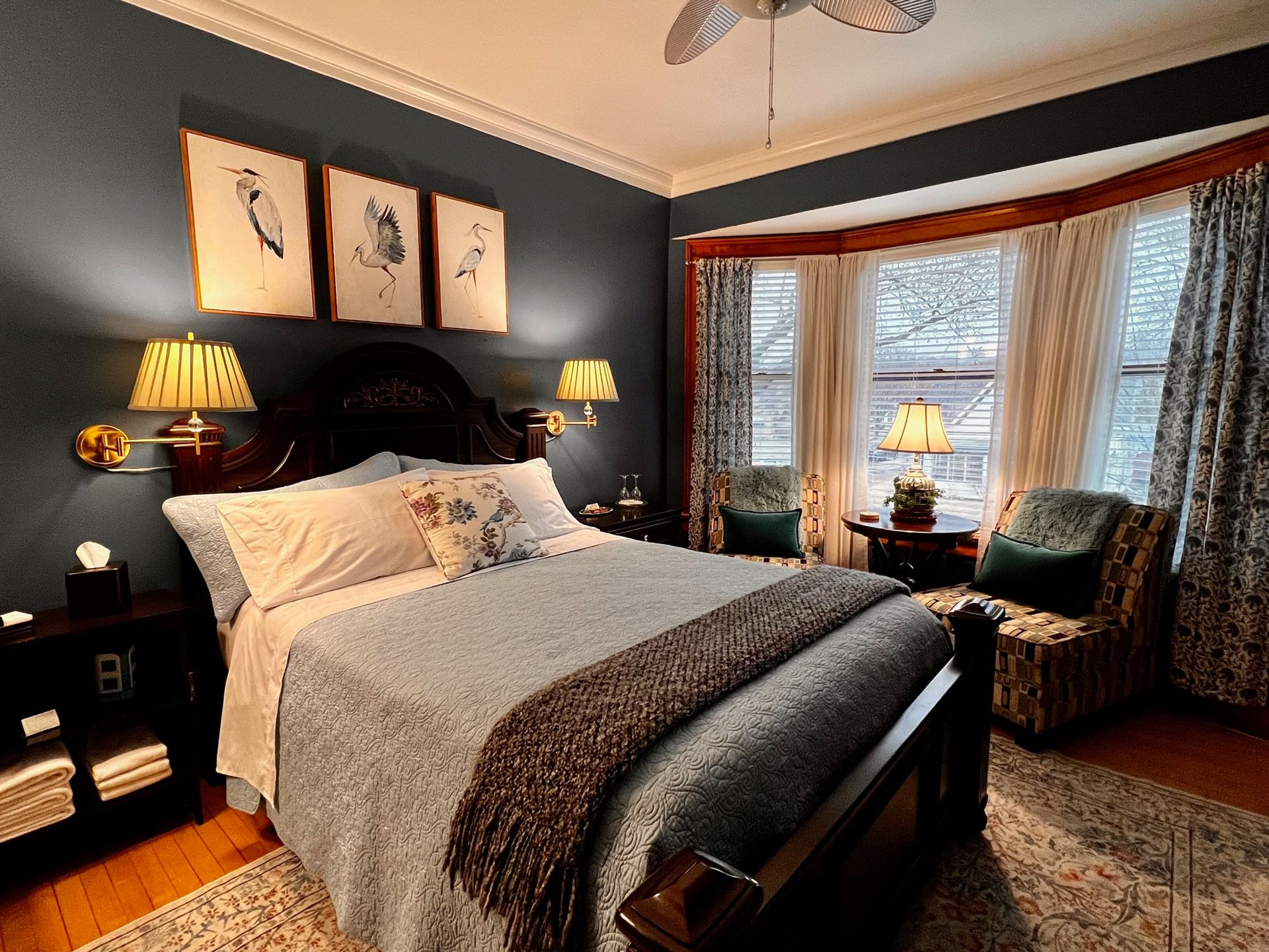 Cozy bedroom with a dark blue wall, bay window, bed, and two patterned armchairs. Artwork hangs above the bed.