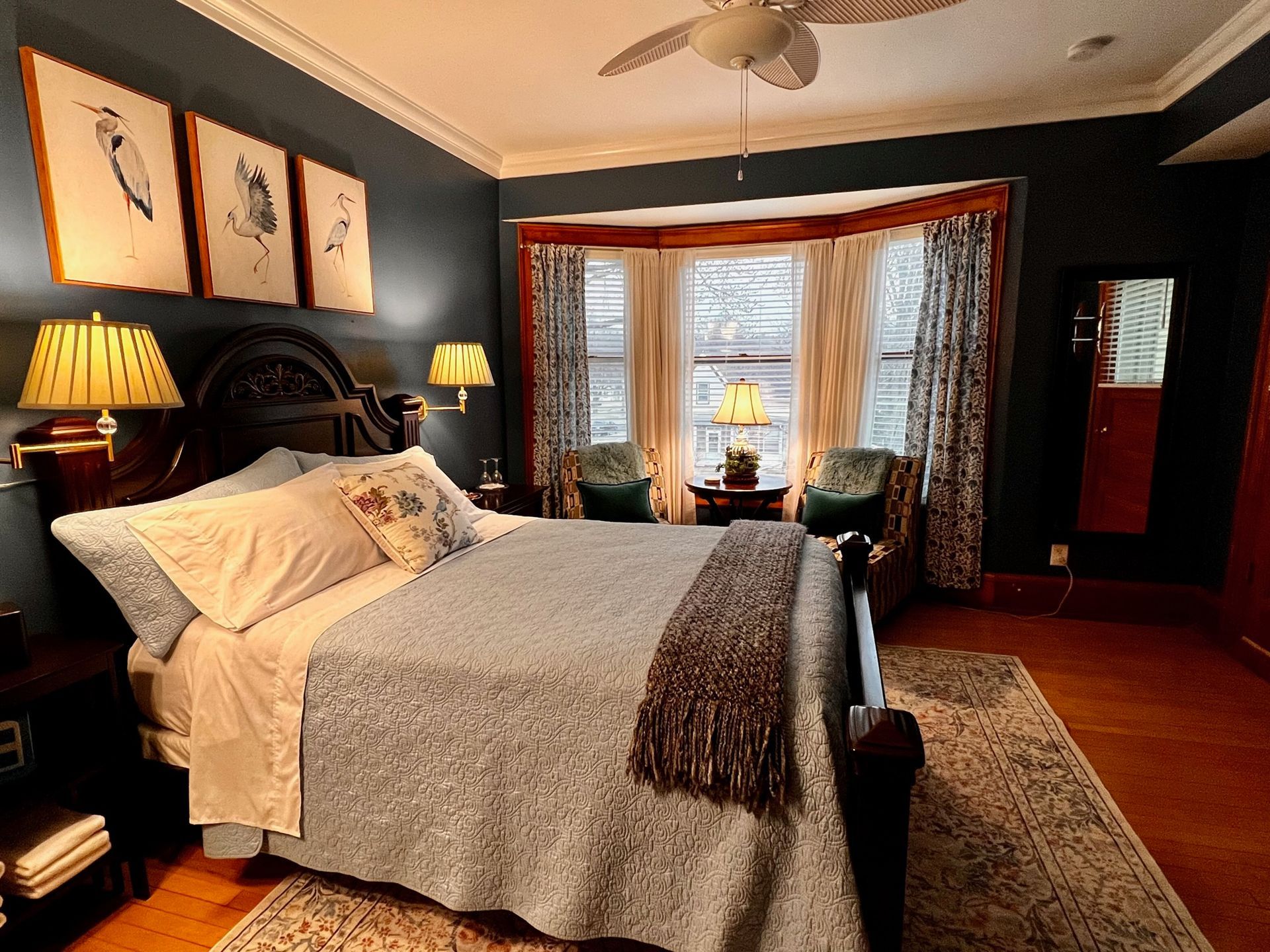 A cozy bedroom with a bed, bay window, and dark blue walls. Artwork and lamps flank the bed.