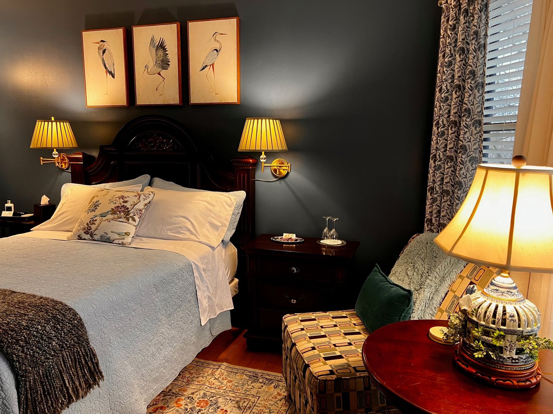 Bedroom with dark blue walls, a bed with light blue quilt, and decorative bird artwork. Two bedside lamps provide warm light.