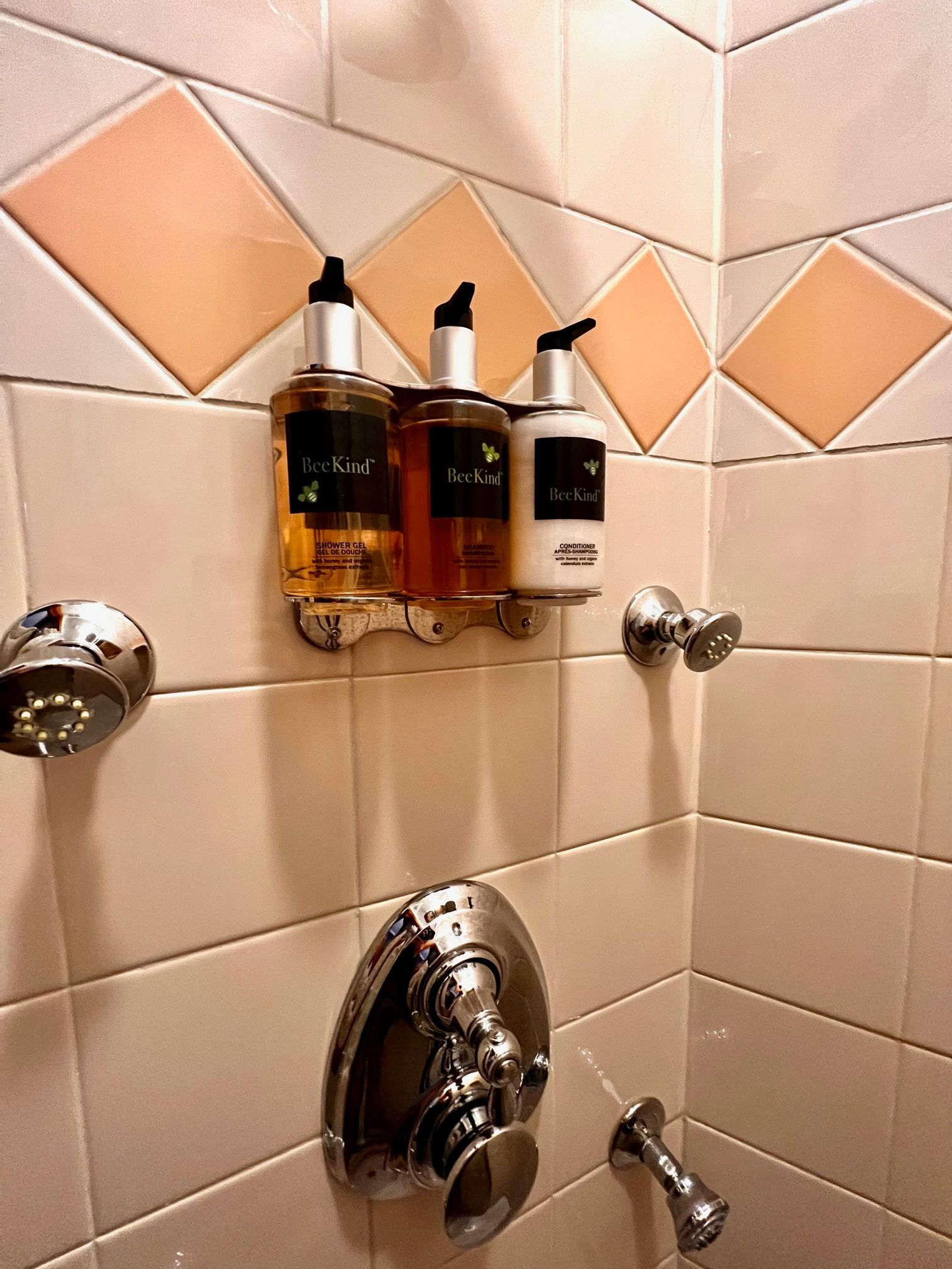 Shower with shampoo and soap dispensers mounted on a tiled wall. Pink and white tiles with chrome fixtures.