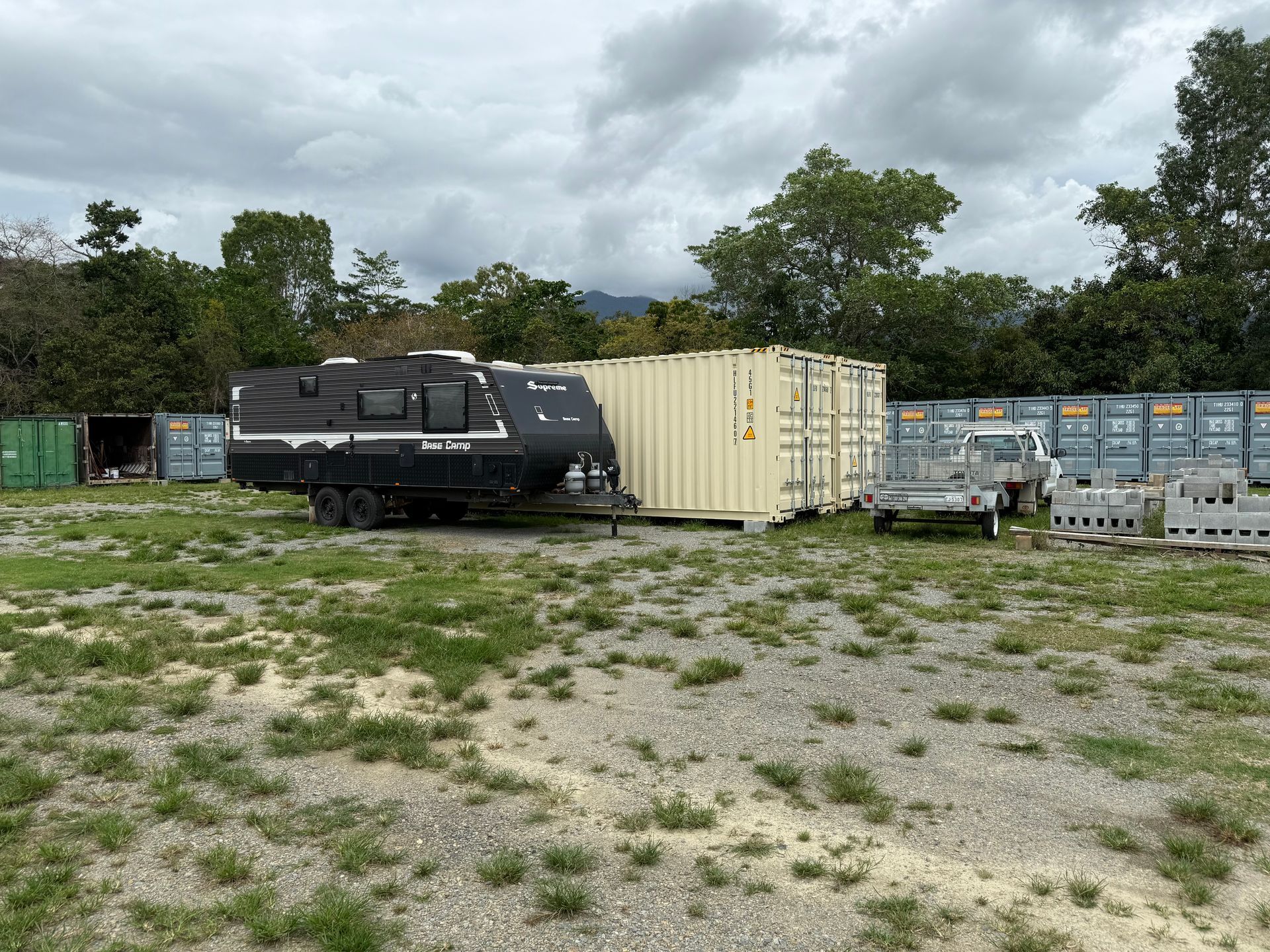 A dark travel trailer parked next to a beige shipping container on a grassy lot with other containers and trees  — Cairns Budget Storage In Mount Sheridan, QLD