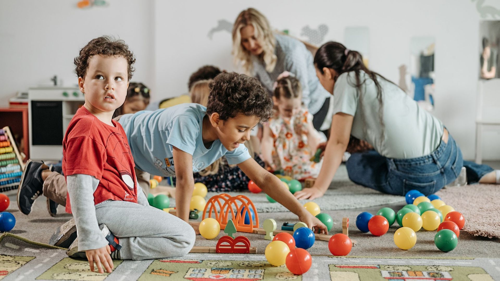 Children playing with balls and toys on a rug in a playroom, with adults supervising.