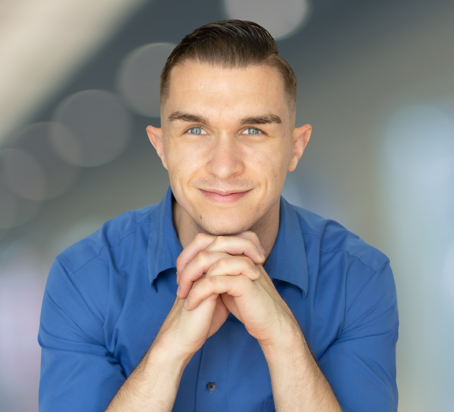 Man in a blue shirt sitting with hands clasped, smiling against a blurred gray background
