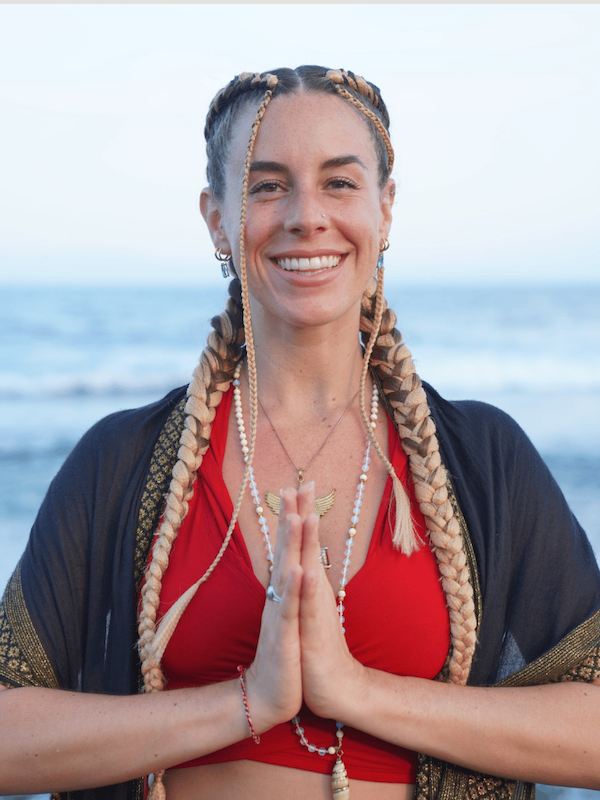 Smiling woman with braided hair and prayer hands on a beach at sunset, wearing a red top and shawl