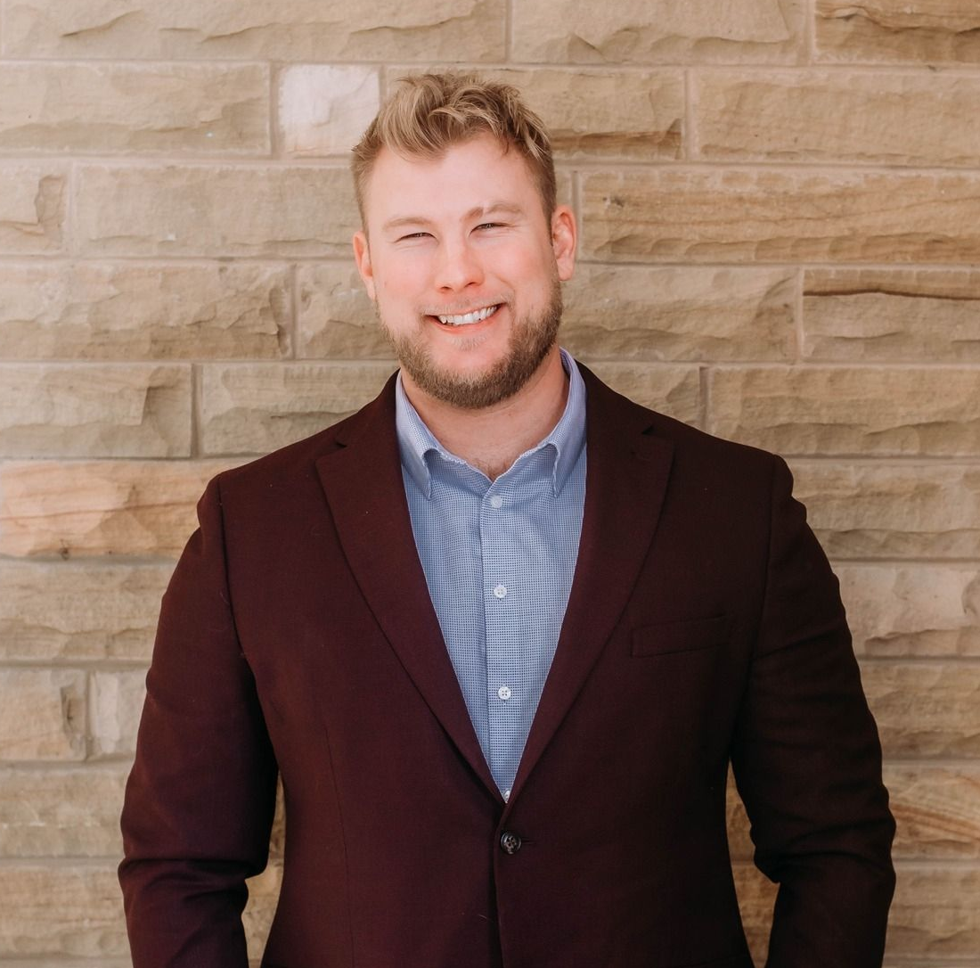 Smiling man in a burgundy blazer and blue shirt standing against a stone wall with hands in pockets