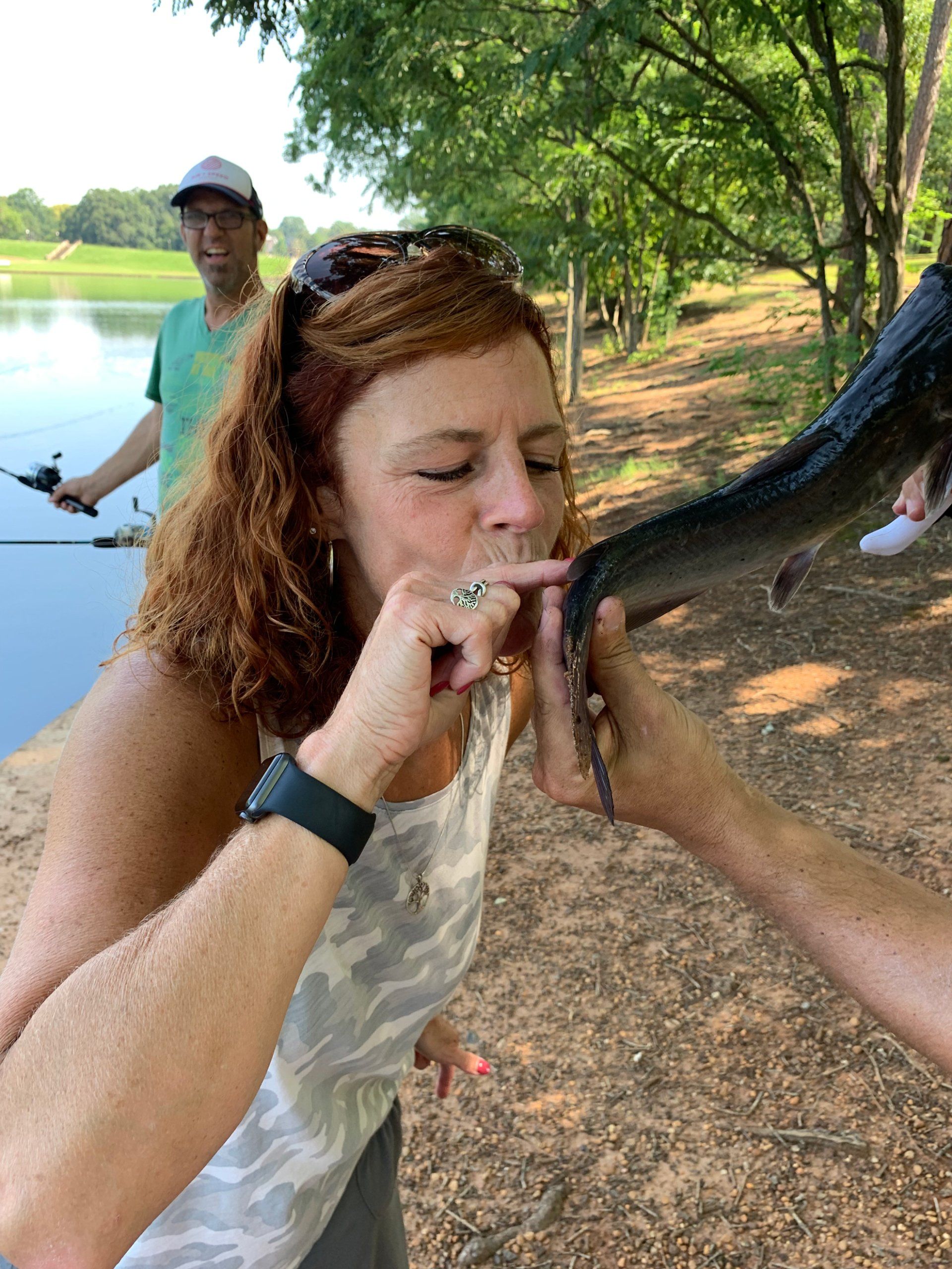 A woman is biting a fish while a man is fishing in the background.