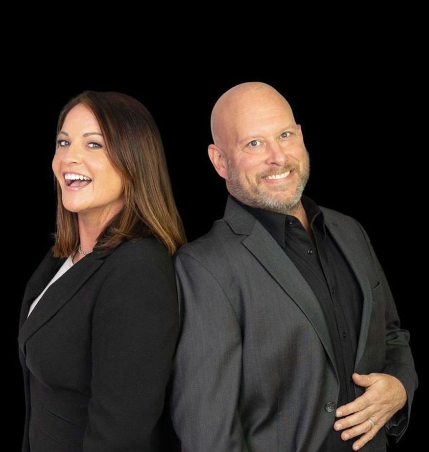 Two people in black suits posing back-to-back against a black background, smiling