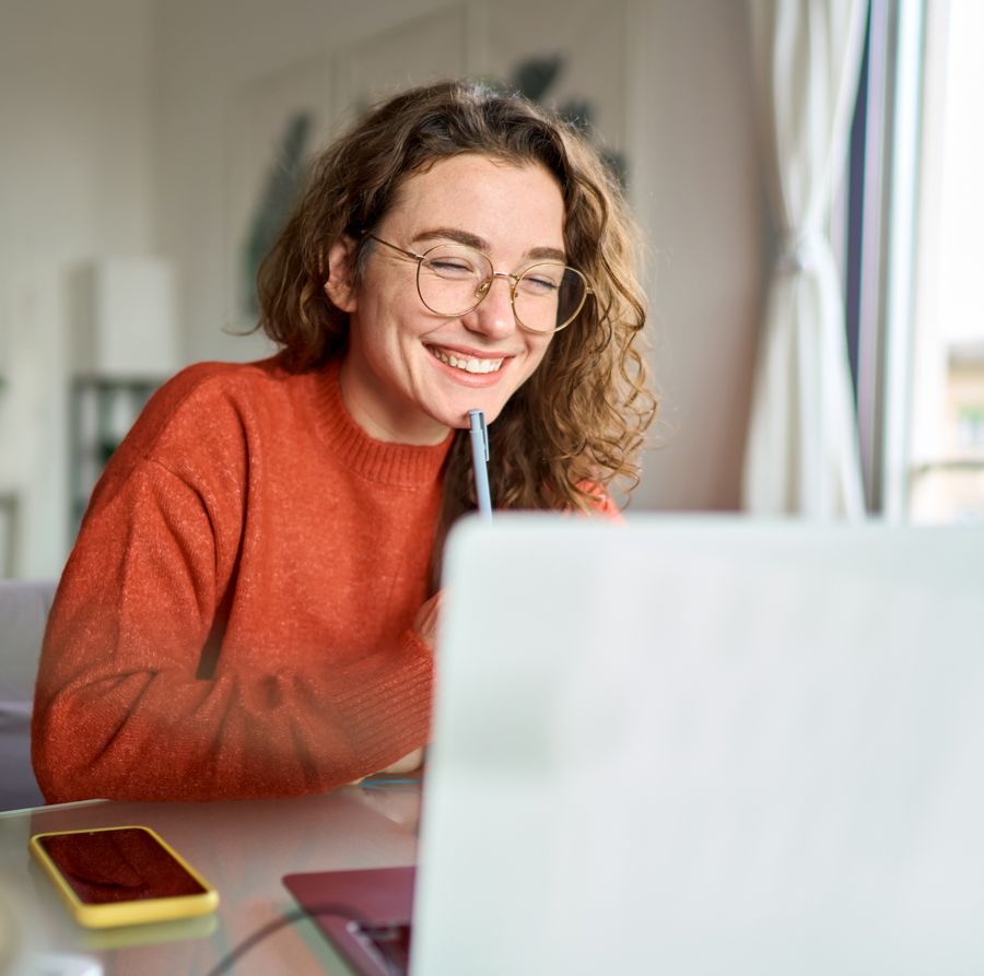 Smiling person in an orange sweater using a laptop at a desk near a window