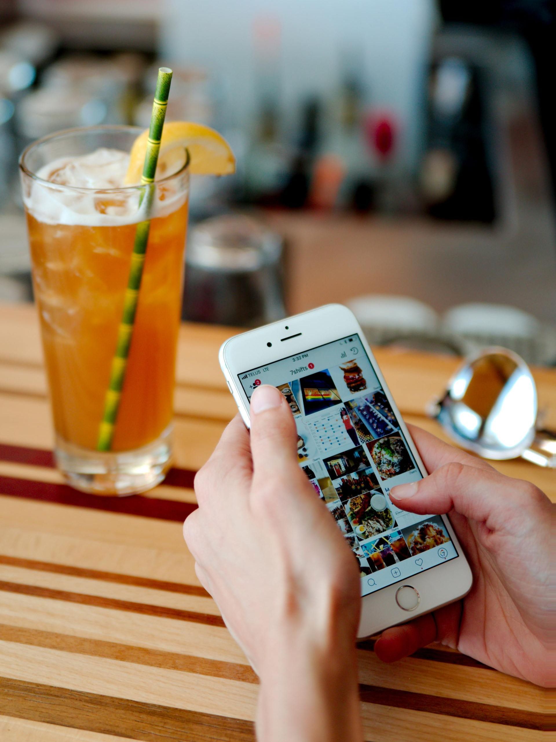 Hands holding a smartphone displaying a photo grid app, with a tall iced drink and sunglasses on a striped wood table.