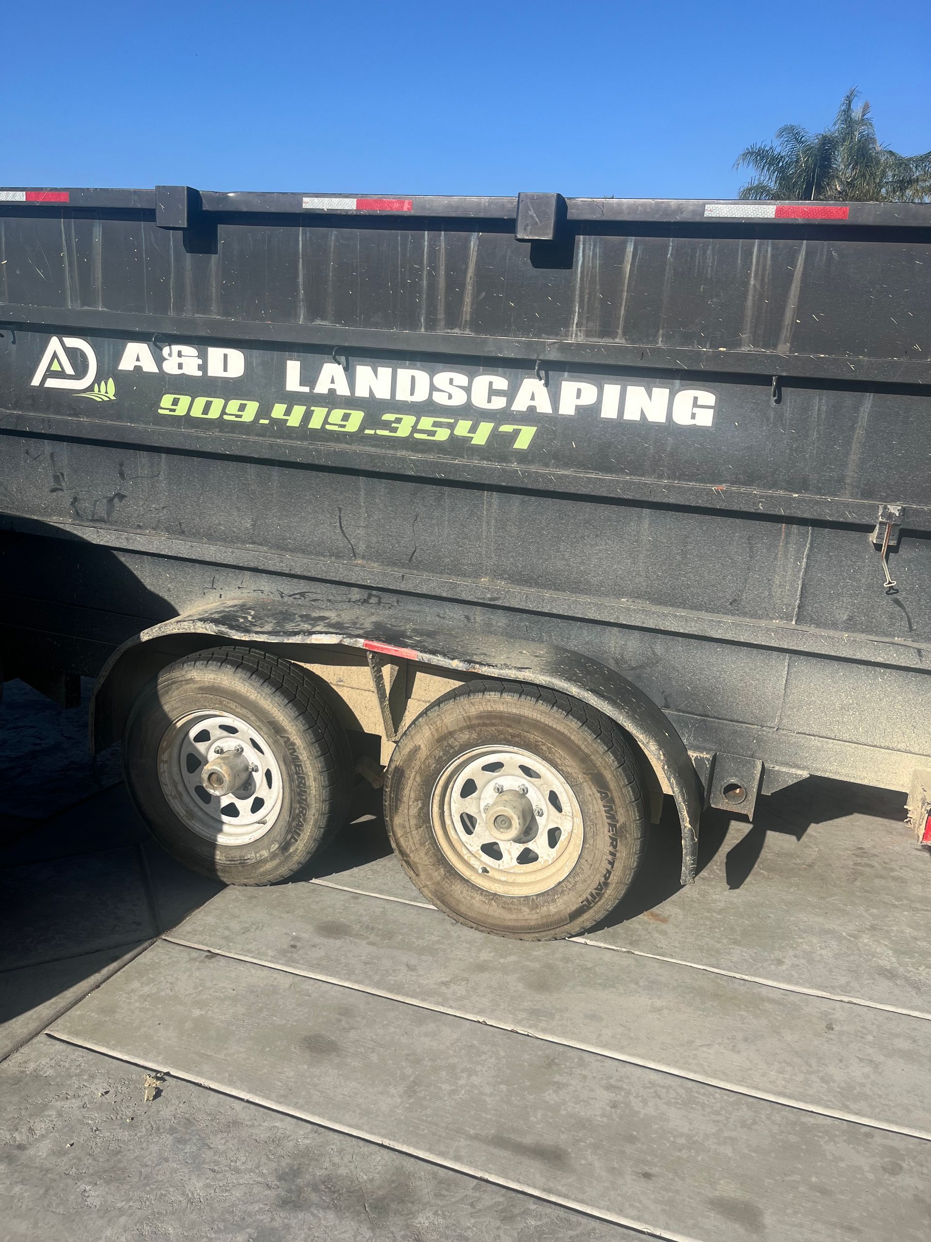 Black trailer with A&D LANDSCAPING logo and a phone number, two dirty tires on asphalt, bright sky.