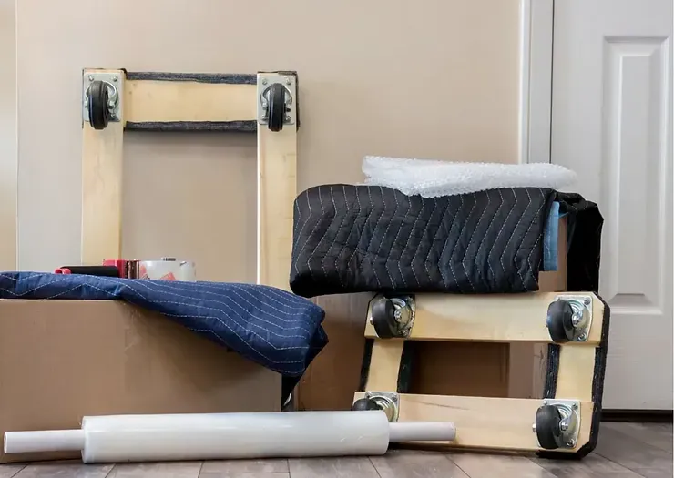 Moving supplies: cardboard box, dollies, packing blankets, tape, bubble wrap, and plastic wrap.