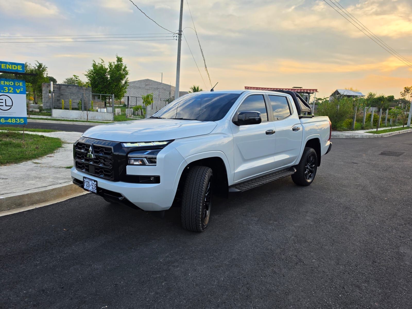Camioneta blanca estacionada en una calle al atardecer con casas y árboles al fondo.