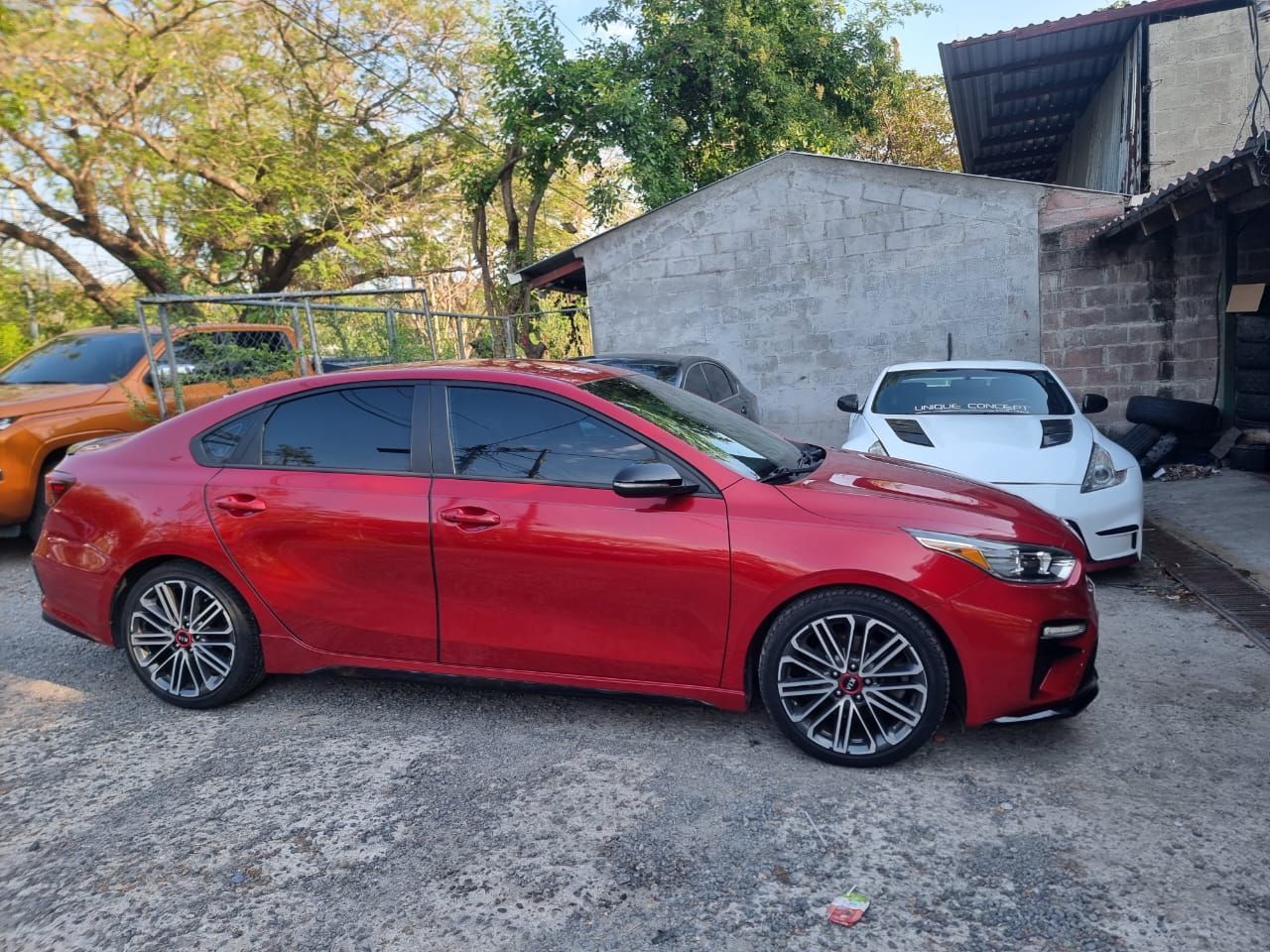 Un sedán rojo estacionado en un terreno de grava junto a un coche blanco y un pequeño edificio, con árboles al fondo.
