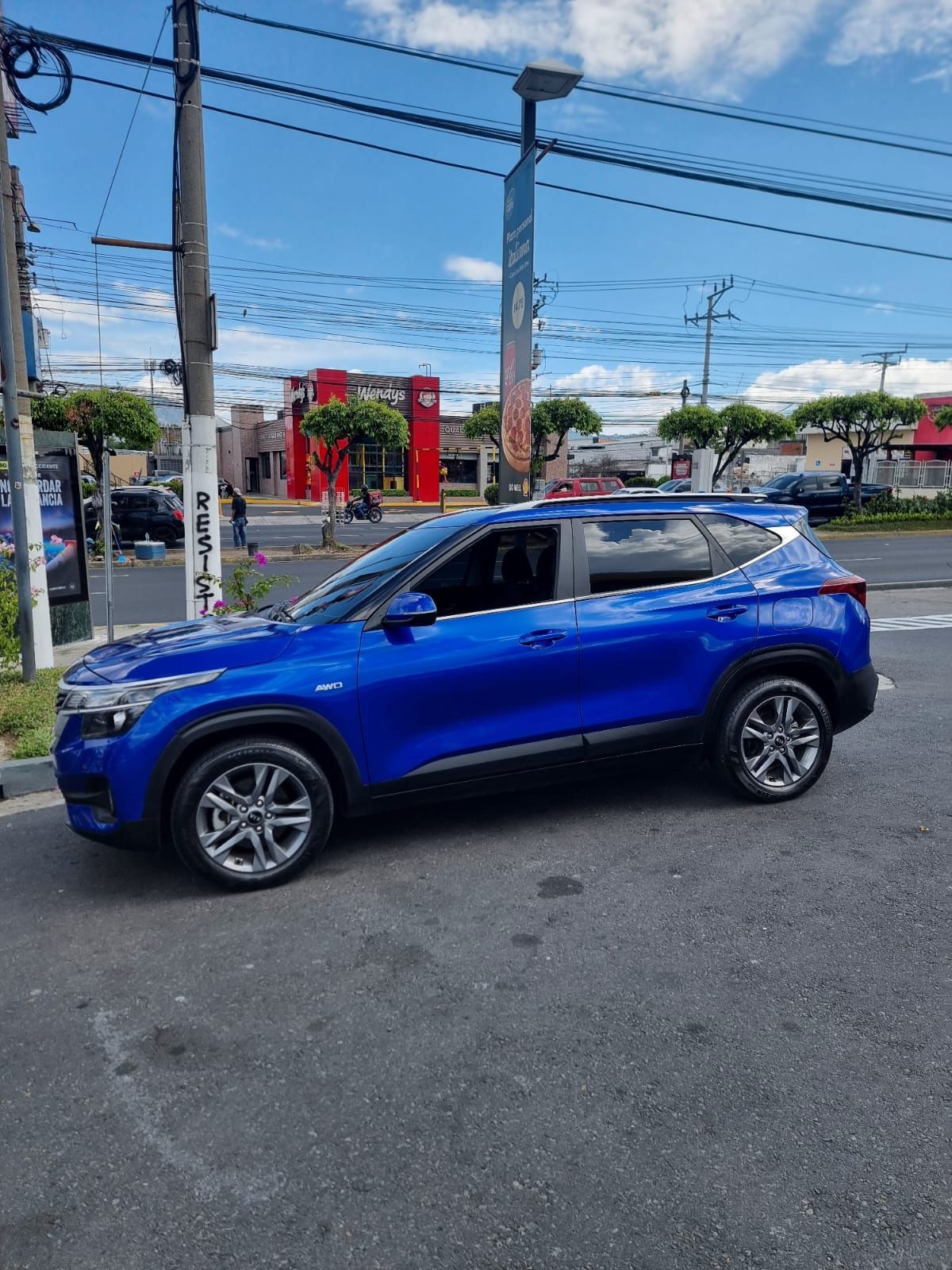 Un SUV azul estacionado en una calle bajo un cielo nublado, con líneas eléctricas y edificios al fondo.