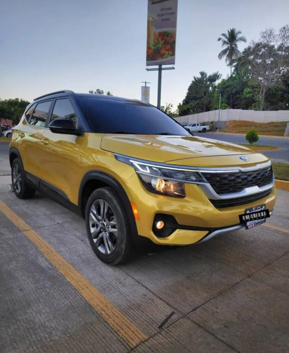 Un SUV amarillo estacionado al costado de la carretera bajo un cielo nublado, con palmeras y un letrero al fondo.