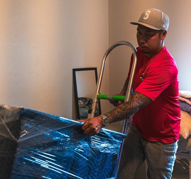 Man in red shirt and cap using a hand truck to move a wrapped piece of furniture indoors.