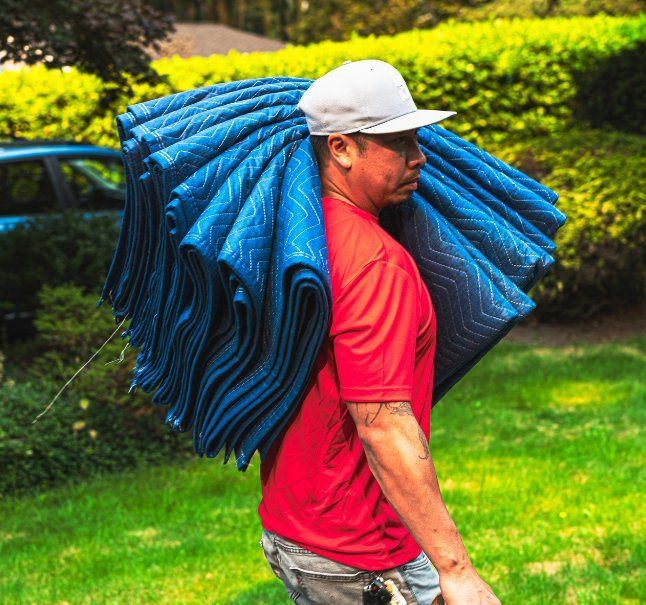 Affordable Movers' team member in red shirt carries a stack of blue moving blankets outdoors.
