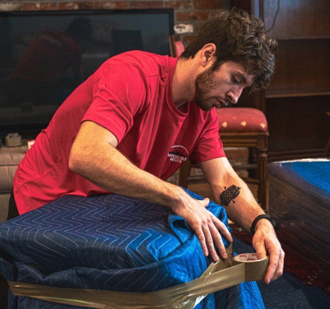 Man in red shirt taping padding around furniture. Indoors, focused expression.