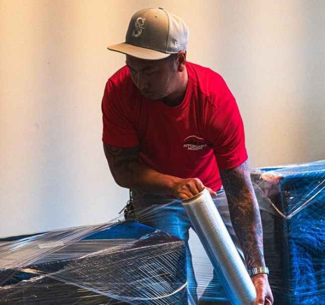 Man in red shirt and gray cap wrapping furniture in plastic wrap.
