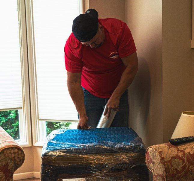 Man in red shirt wrapping furniture with plastic wrap. Window and chairs in the background.