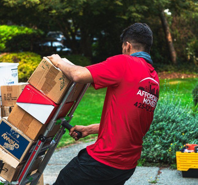 Man in red shirt uses a dolly to move stacked boxes outdoors.