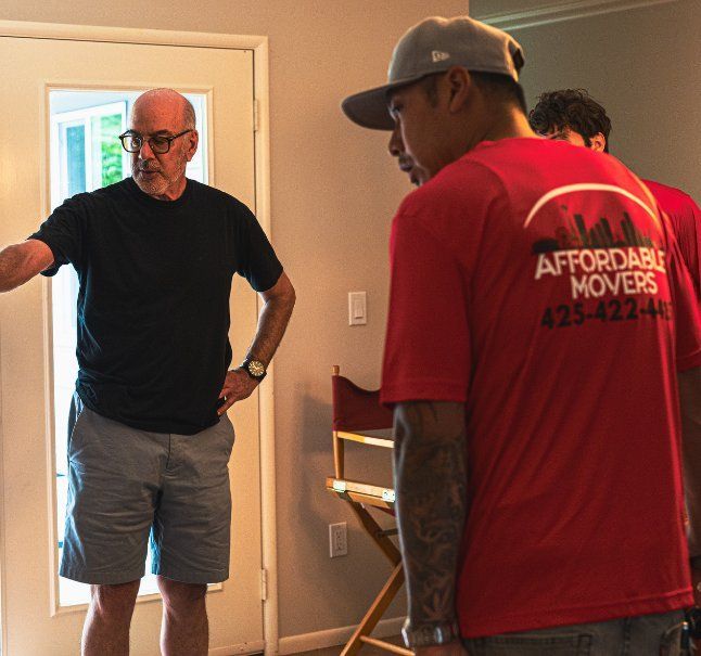 Man directing movers inside a home. Movers wear red shirts, door and window visible.