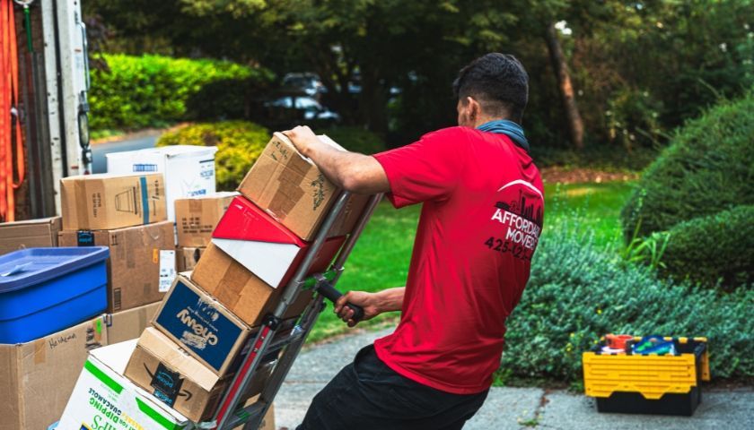 Person in red shirt using a hand truck to move boxes outside a house; moving day.