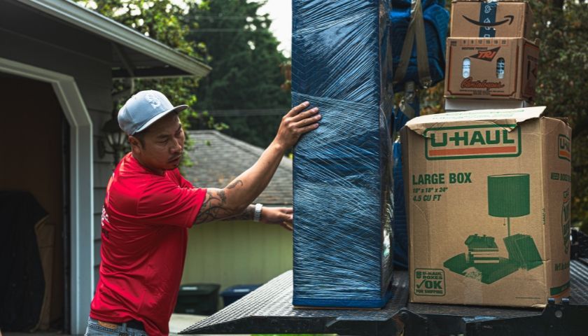 Man in red shirt loads a wrapped furniture item onto a U-Haul truck next to cardboard boxes outside a garage.