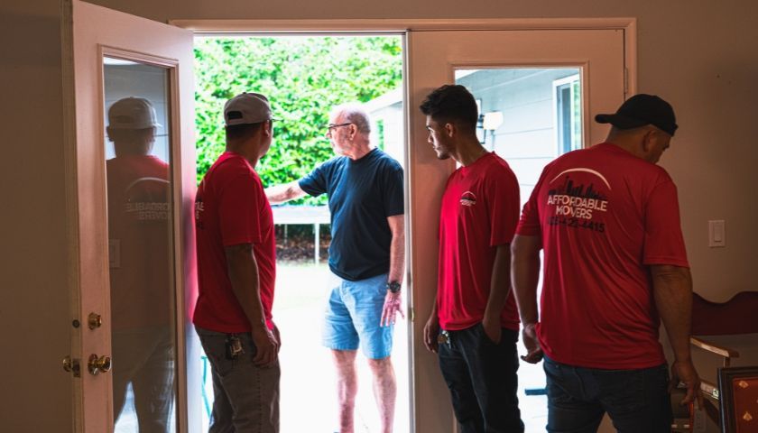 Four movers in red shirts and a man in a blue shirt and shorts in a doorway, likely discussing a move.