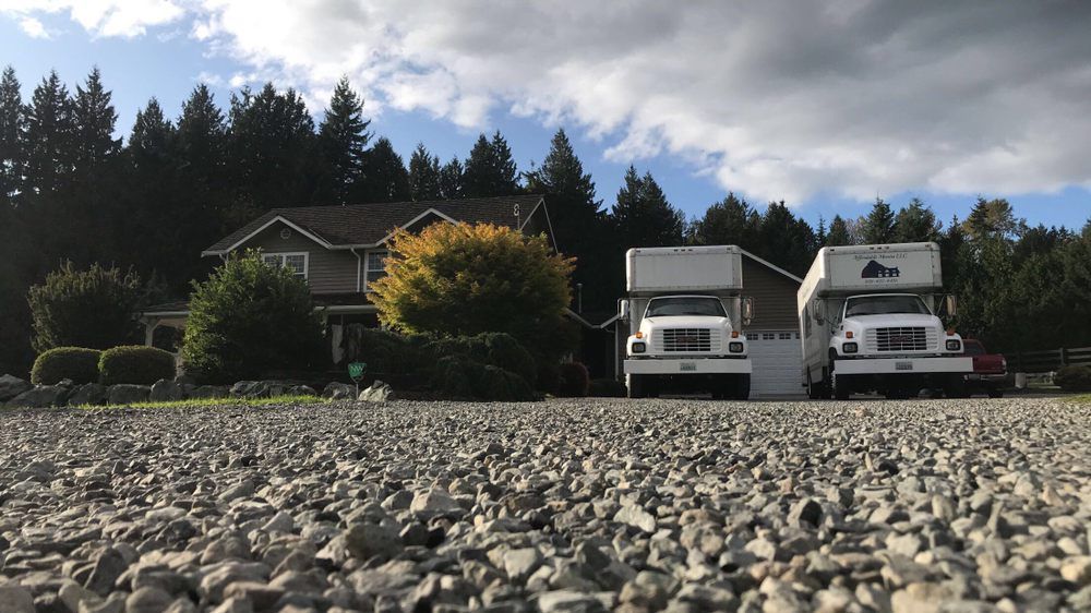 Two moving trucks parked in front of a house on a gravel driveway, trees in the background under a blue sky.