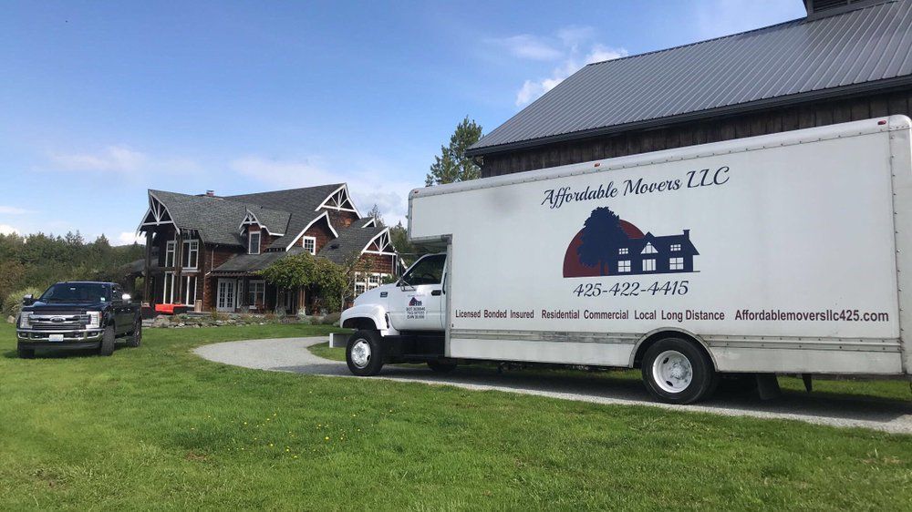 Moving truck parked in front of a large house, with a truck parked in the yard on a sunny day.