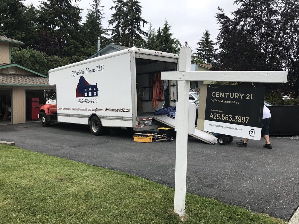 Moving truck at a home with a Century 21 sign. Loading and unloading, driveway, trees, and overcast sky.