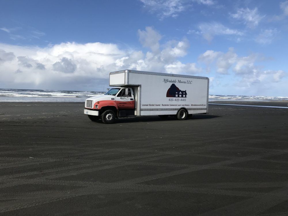 White moving truck on a black sand beach under a partly cloudy blue sky.