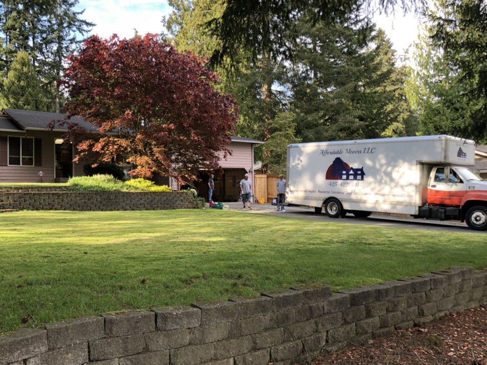 Moving truck parked in front of a house. People loading items. Green lawn, trees, sunny.