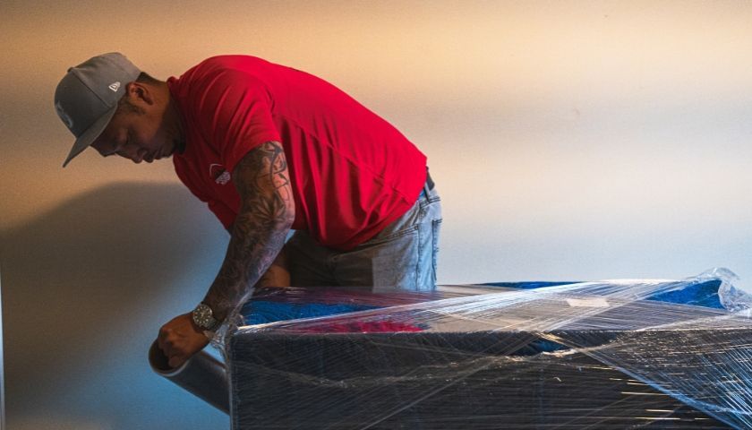 Man in red shirt and gray hat wrapping a blue object with clear plastic wrap.