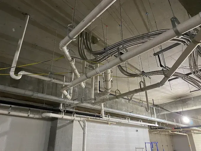 Overhead view of unfinished construction, showing pipes and electrical conduits attached to a concrete ceiling.