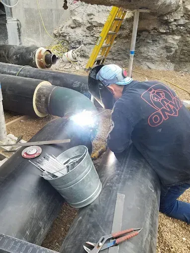 Welder working on insulated black pipes, using a welding torch outdoors.